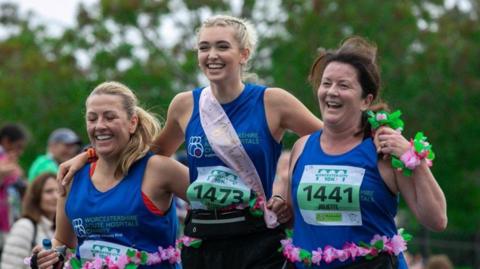 Three women who took part and are wearing blue are arm in arm and smiling at the camera. Other people and trees in the background are blurred.