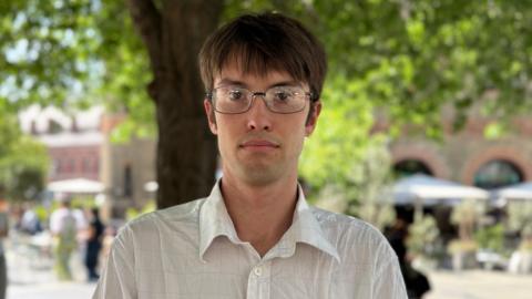 Paddy Friend in a white shirt with a thin check pattern. He has brown hair and glasses.
