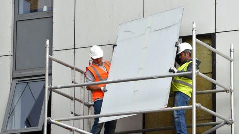 Two workmen in white hardhats and hi-vis vests removing external cladding from Burnham Tower on the Chalcots Estate in Camden in 2017