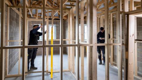 A stock picture of two men working in a house that is partly built with wooden beams put up to divide future rooms.