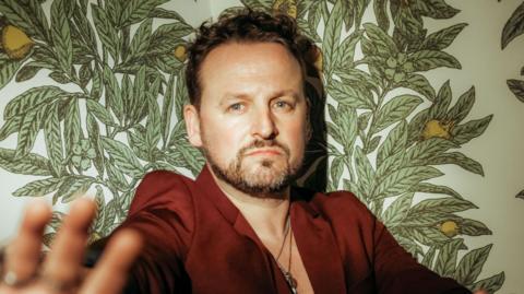 Vince Freeman in a red jacket standing against a wall with a lemon tree on him