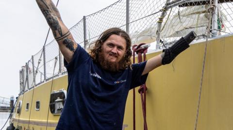 A man with long curly brown hair lifts his arms into the air. He is wearing a blue T-shirt and standing next to a yellow boat. His left arm has no hand.