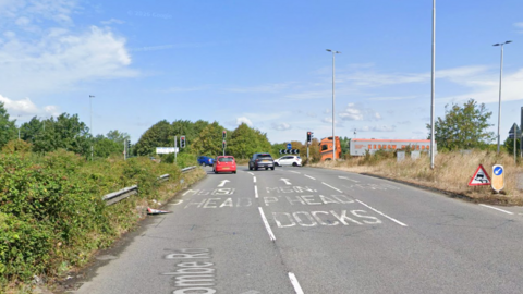 A motorway junction on a sunny day. Cars are queuing at traffic lights. Signs on the road read P'head and P'head Docks.