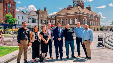 Six men and two women stand in Stockton town centre. The old town hall building is behind them. It is sunny