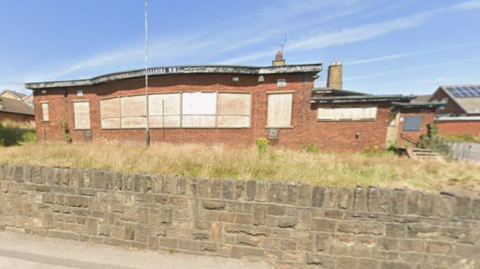 A single-story, redbrick building with boarded-up windows.