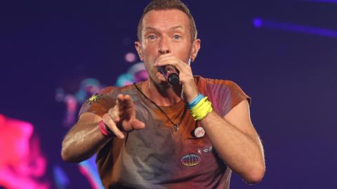 Chris Martin, wearing a multi-coloured T-shirt and wristbands, performs on stage at Wembley Stadium in 2022