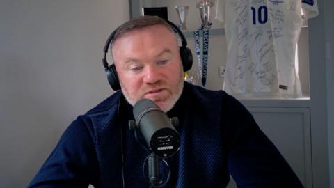 Wayne Rooney talks into a microphone while wearing headphones. An England shirt is on display behind him