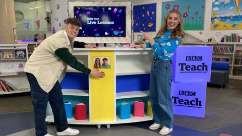 Presenters Joe Tasker and Maddie Moate on the set of the World Book Day Live Lesson. Maddie is pointing towards a TV screen that has a BBC Teach Live Lessons graphic on. The console has a large green button that Joe is pressing.