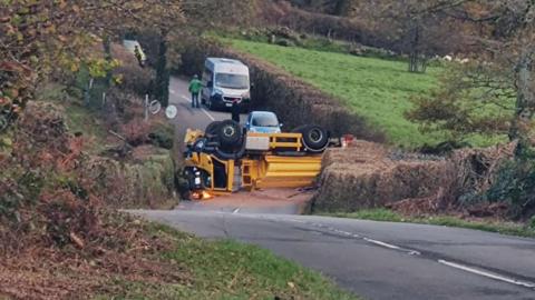 A yellow gritting lorry on its roof with road salt spilled onto the road after it overturned on a Dartmoor road. A white van and blue car are on the other side of the lorry. Three people including a police officer are walking along the road.