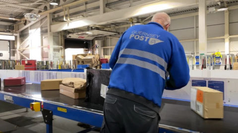 A Guernsey Postal worker in a blue branded top at a conveyor belts of parcels.