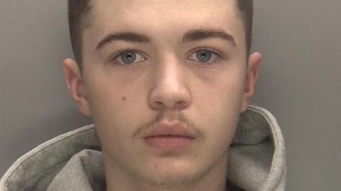 A man with blue eyes and a slight moustache looks into the camera with short dark hair. The hood of a grey hoodie can be seen folded behind his neck.