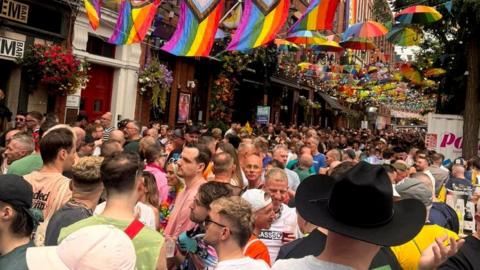 A packed roadway in front of bars in Manchester's Canal Street in the Gay Village. Pride rainbow flags and umbrellas are strung across the street. The tightly packed crowd continues into the distance. 