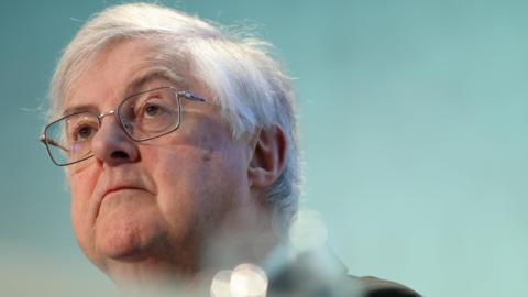 Mark Drakeford, wearing glasses, in front of a pale blue background. He is wearing a dark suit which is slightly out of shot.