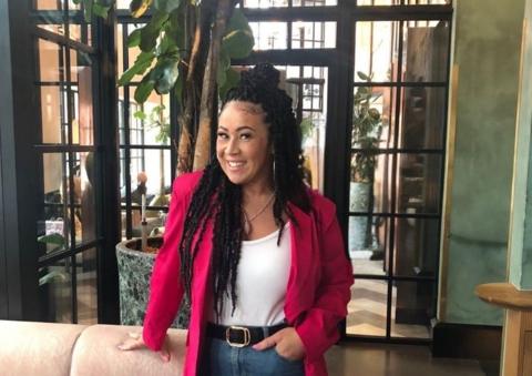 Helena Boyce stands smiling at the camera. Her hair is black and in goddess braids, which have been tied up in a half up, half down style. She has large silver hoop earrings in and wears a white tank top underneath a fuschia pink blazer.