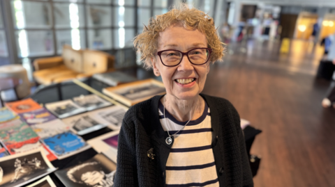 Julia Kirby has short blonde tight curly hair. She has round purple-framed glasses and wears a black and white striped t-shirt and black knitted cardigan. She is smiling and standing next to a piano with posters spread out across the top