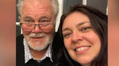 Emma Herrick (right) with long dark brown hair wearing a pale blue top takes a selfie of her and her dad, Roy, who has grey hair and glasses and is wearing a checked shirt and black jumper. They are both smiling.