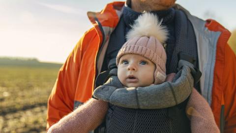 A smiling baby wearing a pink hat is looking out while being carried in a grey baby sling worn by her father, who wears a thick blue jumper and orange rain jacket.