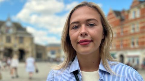 Daisy Blakemore-Creedon has blonde hair and is standing outside in Peterborough city centre in front of the Guildhall building. She is wearing a butter yellow coloured T-shirt with a white and blue striped shirt over the top. She is looking directly at the camera and the sky is blue in the background behind her.