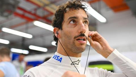 Daniel Ricciardo puts ear protectors in while wearing his fireproof overalls in the pit area