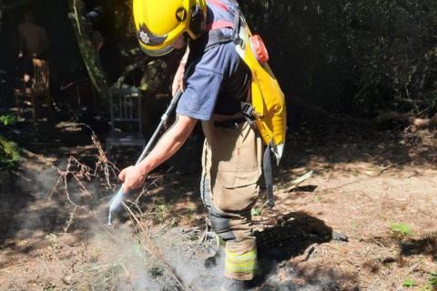 A firefighter with a water hose and backpack extinguishing a small camp fire in a forest. He is wearing the beige firefighters overalls with a navy blue t-shirt and yellow helmet. It's a sunny day and the light is streaming in through the trees.