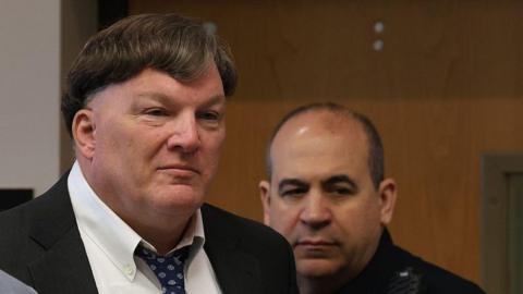 A man in a suit and blue patterned tie looks off to the right as a security guard stand behind him