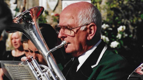 An older man playing a tenor horn with laminated music in front of him. He is wearing a deep green blazer. It is a visibly old image, taken in 2004.