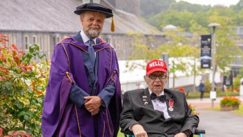 Actor Toby Jones is standing alongside Neil Baldwin. Jones is wearing a purple gown and black cap with gold tassle. Mr Baldwin is sitting in a wheelchair and is wearing a red baseball cap with white letters that read "Nello". He is also wearing his MBE medal and a number of pin badges.