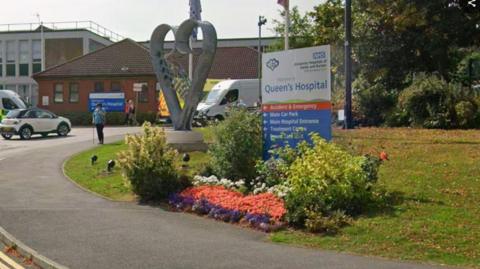 Sign reading 'Queen's Hospita' on grassy verge at the side of a drive running up to a hospital building 