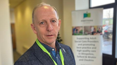 Grey-haired man wearing blue shirt, blue blazer and green lanyard, posing for a photo, with how company's pull up banner behind him.