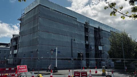 This image shows a large multi‑storey building completely wrapped in grey protective sheeting, with scaffolding visible along one side. The covering stretches from ground level to the roof. Several layers of scaffolding are exposed on the right‑hand section, with metal poles, platforms and ladders visible through gaps in the sheeting.