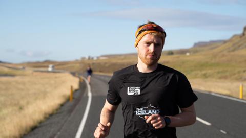 Seb Key running in Iceland - he is wearing a black t-shirt and a headband. He is running on a road, with yellow-ish fields each side.