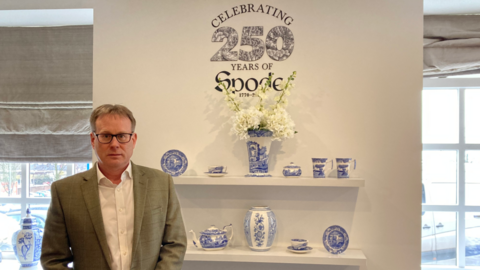 A man in glasses, wearing a suit jacket over an open necked shirt stands next to a display of blue and white decorated pottery. A sign above the pottery says celebrating 250 years of Spode