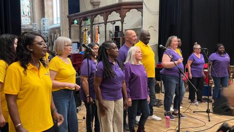 Thirteen members of a choir standing on a stage behind a number of mics. They are stood in a church like hall.