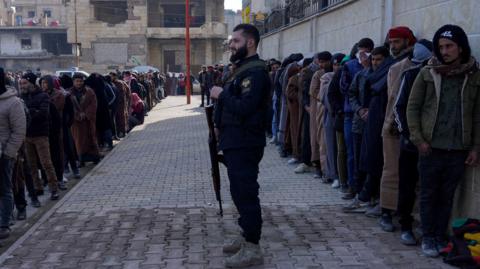A queue of men on the right against the wall of a building, and a queue of men on the left on the edge of a pavement. A man holding a gun pointed to the ground is standing on the pavement. it is outdoors.