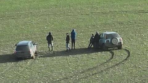 Group of people in a field, next to two cars