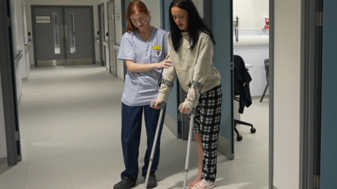 The picture shows a hospital corridor inside a clinical building. A member of hospital staff in scrubs is supporting a young patient as they carefully move forward using a pair of crutches. The patient is dressed casually and wearing slippers