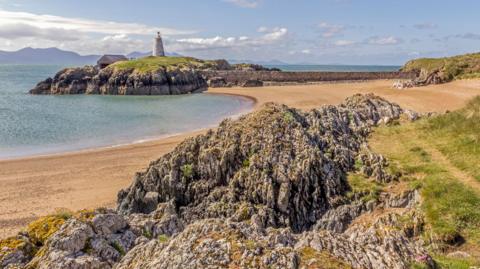 Image shows rocky terrain and a sandy beach in the foreground.
A small island in the background, topped with a lighthouse and an adjacent building.
The island is surrounded by the sea, with mountains visible in the distance.
The sky is mix of blue sky and some clouds above the mountain's on the horizon.