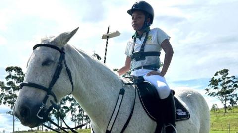 Khosi Dlomo, dressed in white South Africa riding gear and black helmet, sits aloft her white horse Yogi, holding a lance up in her right hand on which a tent pegging target is speared