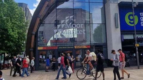 Glass fronted building with The Moor Market written on the glass in white. There are shoppers in front in a pedestrian area.