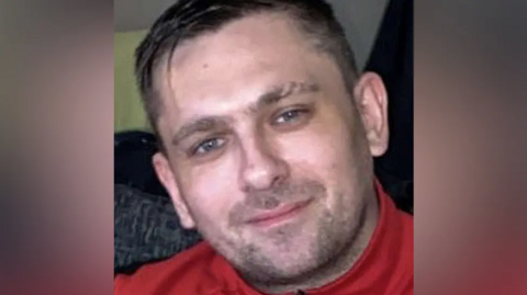 Sean Dock, a man in his 30s with short dark hair and stubble, smiles at the camera. He is wearing a red polo neck.