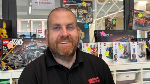 There is a man with a shaved head and a ginger beard, with some grey hairs in there. He is wearing a black polo with 'Brick and Beyond' in red letters on. Behind him is a collection of lego sets, including a Pikachu, football players and a Star Wars set