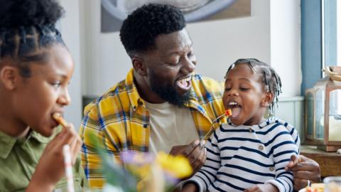 A father sits at the dinner table with two children. The youngest is on his lap and he's feeding them some food.