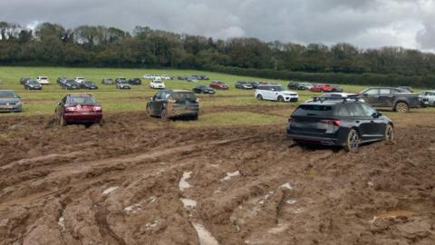 A field covered in wet mud with dep track marks from car tyres. Some of the cars in the foreground are covered in mud while others are parked in the distance.