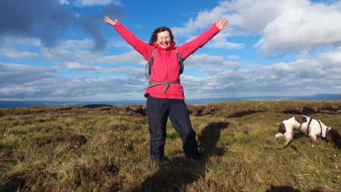 A woman in a red jacket is standing with her arms outstretched to celebrate reaching another summit. The sky is very blue with white clouds and a brown and white dog is next to her investigating a rucksack