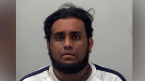 Oaveed Rahman is wearing a white top with a black stripe along both shoulders. He has a beard and he is looking at the camera. It is a custody photo.