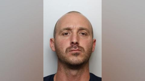 A police mugshot of Sean Deering - a man in his 30s with his shaved hair and stubble. He has brown eyes and is looking into the camera.