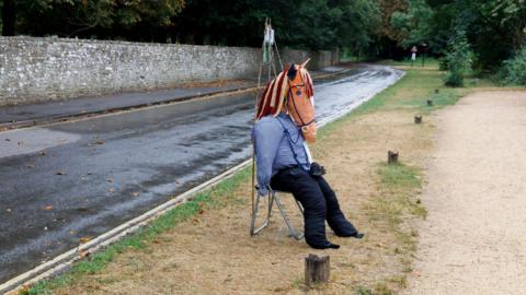A scarecrow with a horse's head on a stool. The stool is on a grass verge at the side of a road. A long stone wall and trees can be seen in the background. 