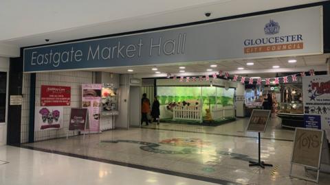 Image of Eastgate Market Hall in Gloucester. There is a large blue and white sign spanning the width of the market entrance. Multiple stalls can be seen inside.