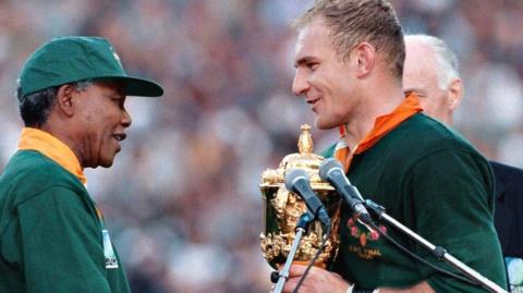 Nelson Mandela presents Francois Pienaar with the Rugby World Cup trophy in 1995, both wearing Springbok jerseys