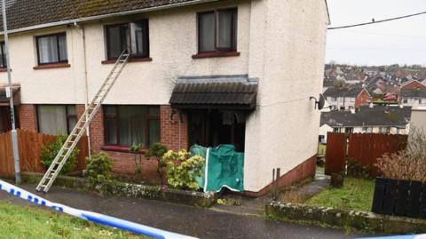 A cream and red brick semi-detached house which sits on a hill, with loads of houses behind. Police tape is visible at the forefront of the picture. There is a green cover over the front door, the windows look black and there is a ladder going up into a first floor window.